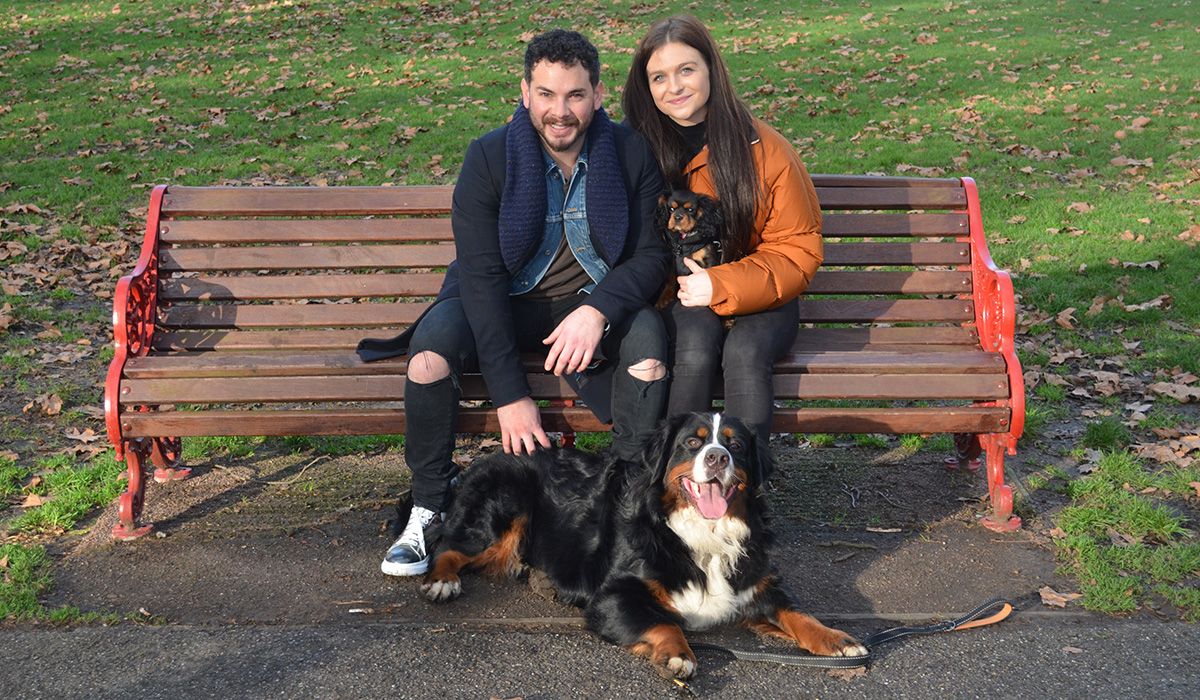 Two people are sitting on a bench. A large black, white and tan dog lies at their feet